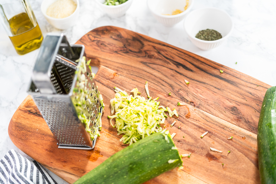 Chopped zucchini and garlic on a wooden board next to a grater and olive oil bottle.