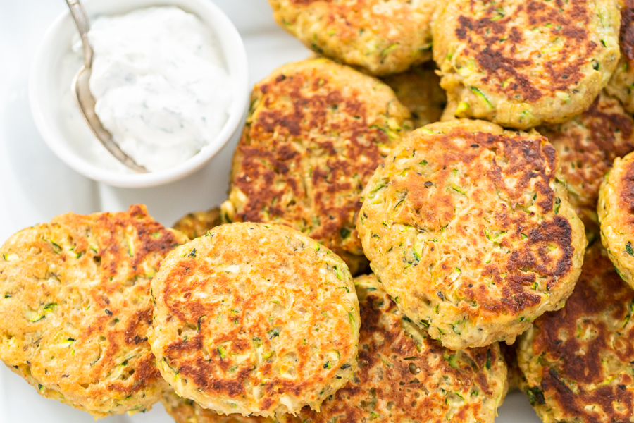 Overhead view of golden-brown zucchini fritters stacked on a plate with dip on the side.