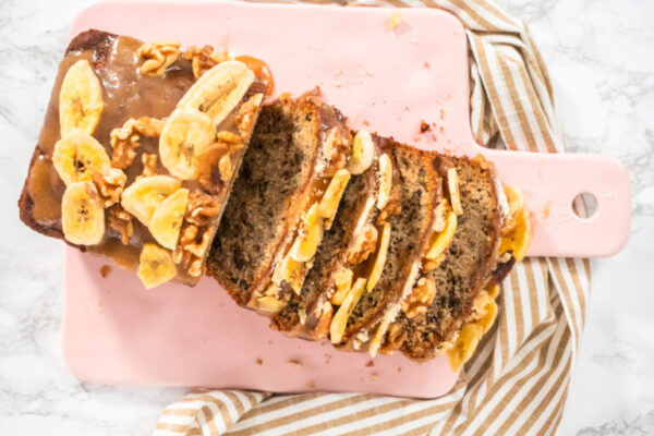 Flat lay. Sliced loaf of banana nut bread drizzled homemade caramel on a pink cutting board.