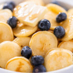 Cooked mini pancakes topped with fresh blueberries in a white bowl – final serving of mini pancake cereal with blueberries and syrup for a sweet breakfast.
