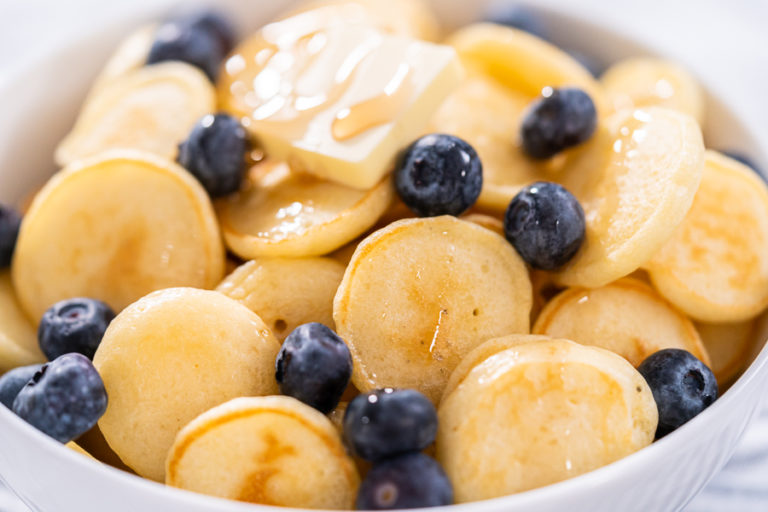 Cooked mini pancakes topped with fresh blueberries in a white bowl – final serving of mini pancake cereal with blueberries and syrup for a sweet breakfast.