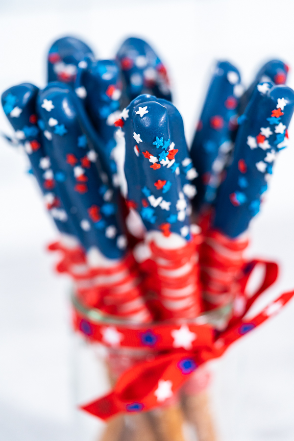 Close-up of red, white, and blue chocolate-covered pretzel rods decorated with patriotic star sprinkles and wrapped in a festive red ribbon, perfect for 4th of July celebrations.