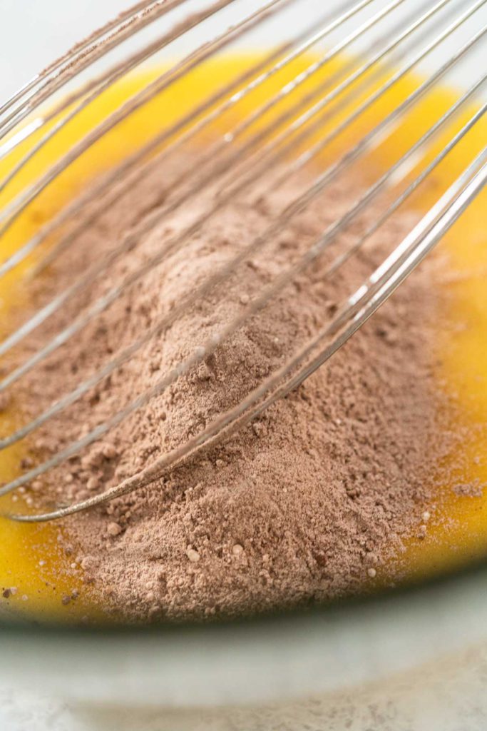Cocoa powder being whisked into a yellow batter mixture in a glass bowl, preparing chocolate cupcake batter.