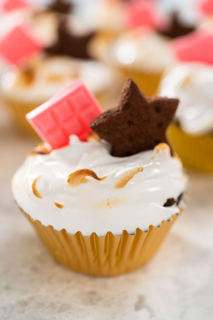 Toasted s’mores cupcake topped with swirled marshmallow meringue frosting, pink chocolate bar, and star-shaped chocolate graham cracker in a gold foil liner.