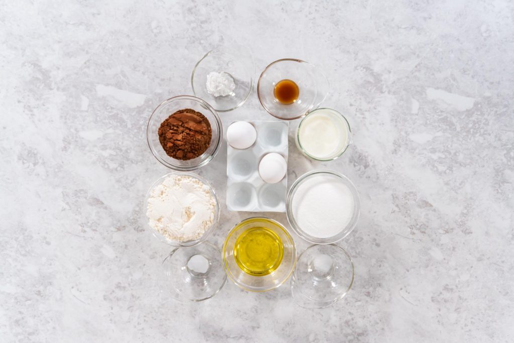 Flat lay of ingredients for chocolate cupcake batter, including eggs, cocoa powder, flour, oil, sugar, vanilla, baking powder, and buttermilk arranged on a light surface.