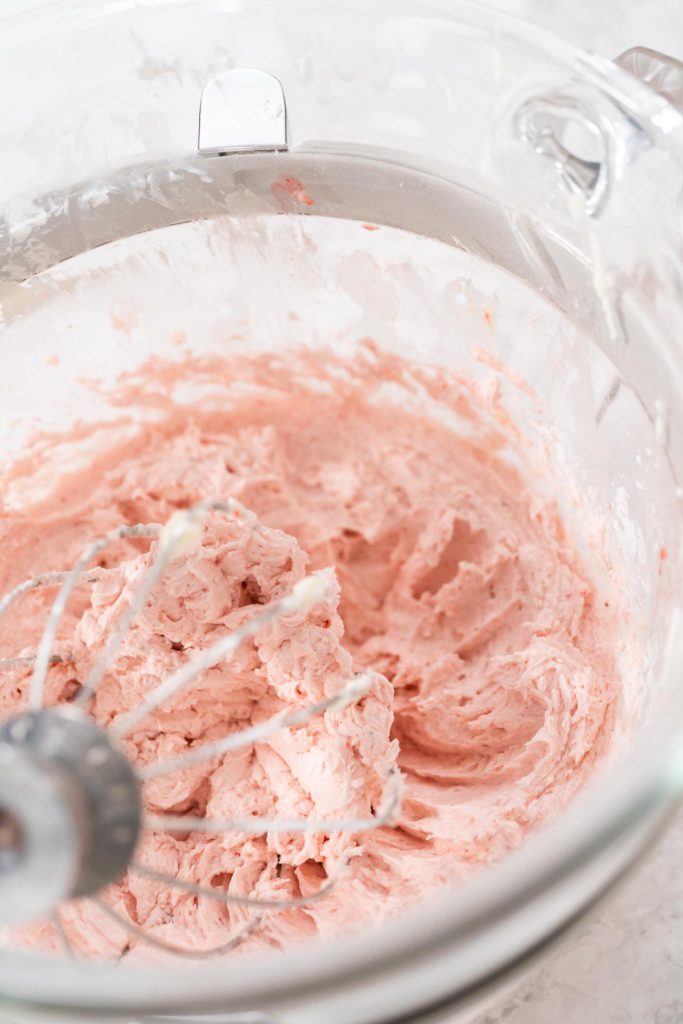 Glass bowl with strawberry frosting mid-whip, attached to a whisk in a stand mixer.