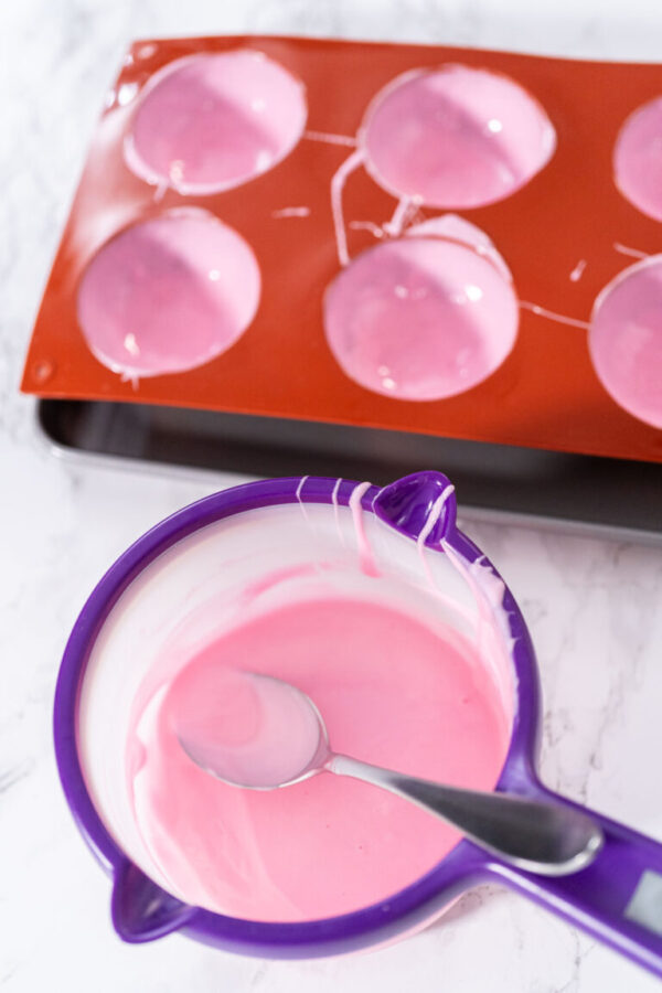 Filling in silicone chocolate molds with melted pink chocolate to prepare hot chocolate bombs.