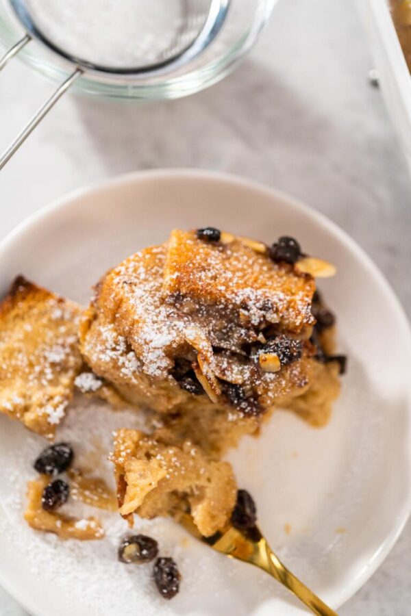 Serving freshly baked bread pudding with raising and almond slivers in a white casserole dish.