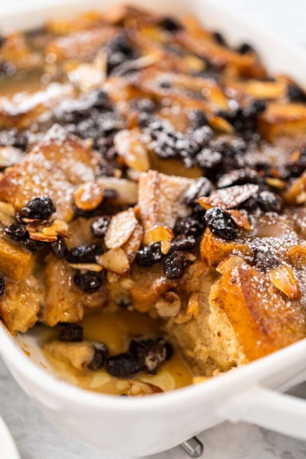 Serving freshly baked bread pudding with raising and almond slivers in a white casserole dish.