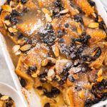 Serving freshly baked bread pudding with raising and almond slivers in a white casserole dish.
