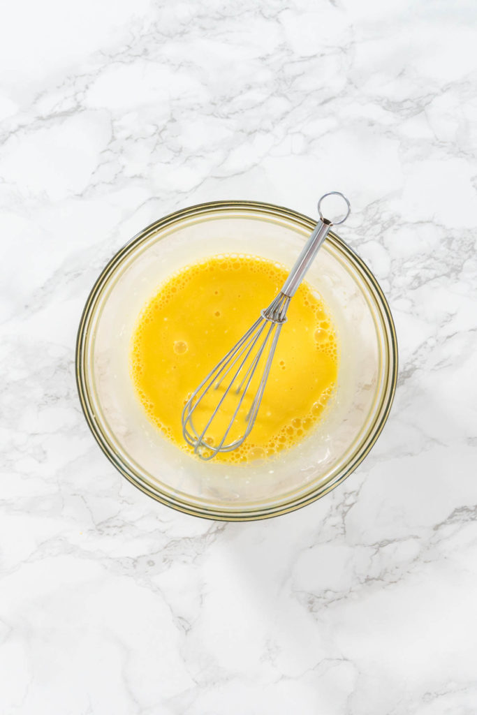 Egg yolks whisked in small bowl on marble counter.