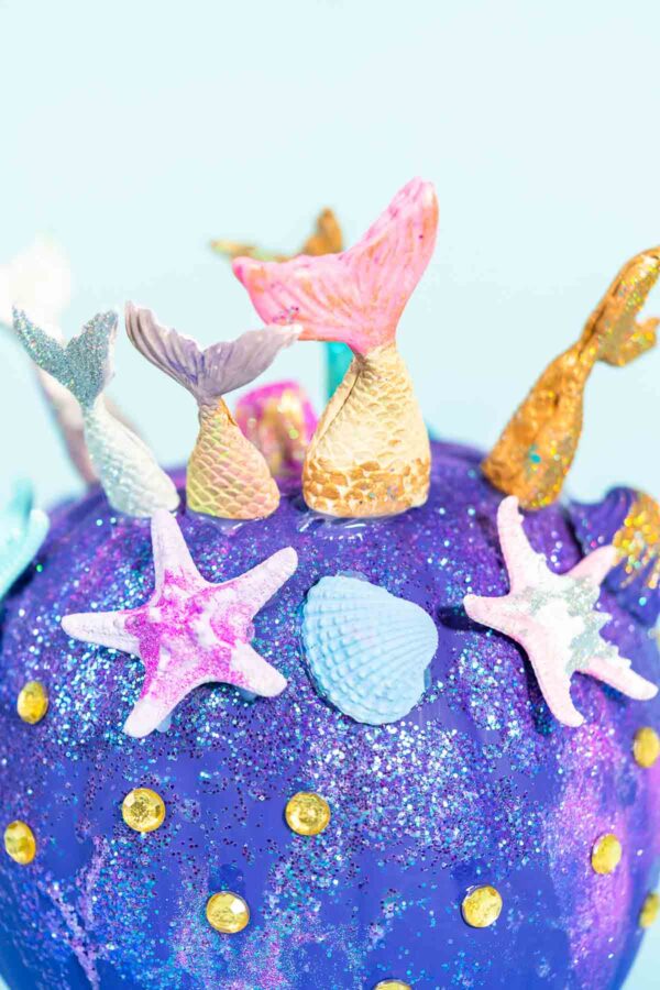 Halloween craft pumpkins decorated with mermaid tails, glitter, and sparkly rhinestones on a blue background.