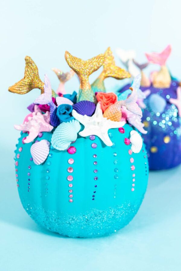 Halloween craft pumpkins decorated with mermaid tails, glitter, and sparkly rhinestones on a blue background.