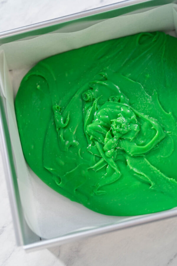 Filling square cheesecake pan lined with parchment paper with fudge mixture to prepare green fudge with red hearts.