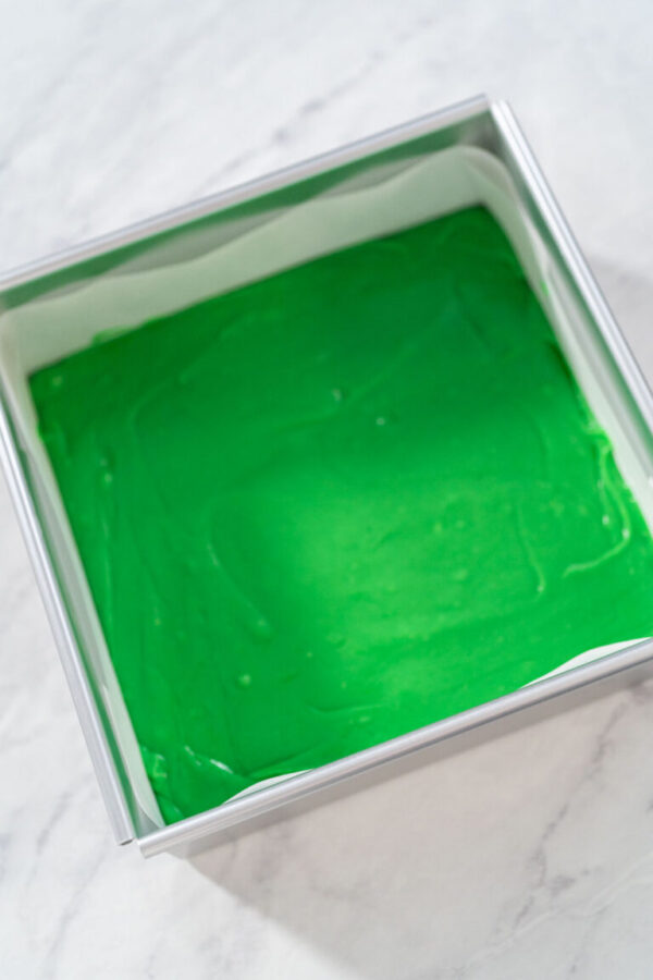 Filling square cheesecake pan lined with parchment paper with fudge mixture to prepare green fudge with red hearts.