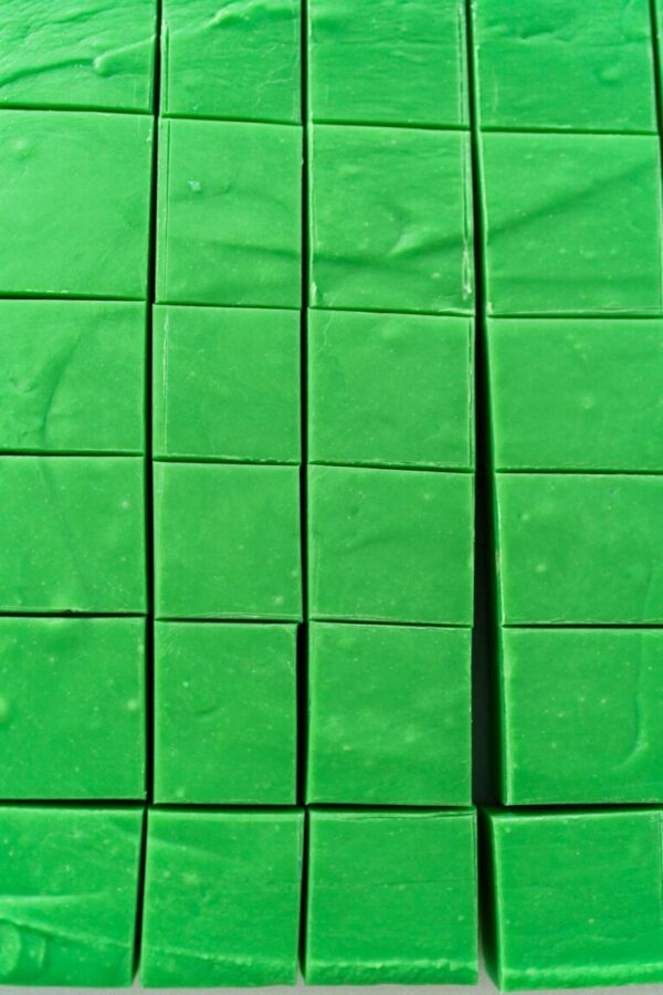 Cutting green fudge with a large kitchen knife into square pieces on a white cutting board.