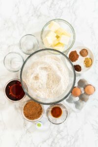 Overhead view of the ingredients laid out for Vibrant Green Gingerbread Cookies for Festive Baking. Ingredients include butter, brown sugar, flour, green food coloring, eggs, cinnamon, cloves, ginger, and maple syrup for sweetness and flavor.