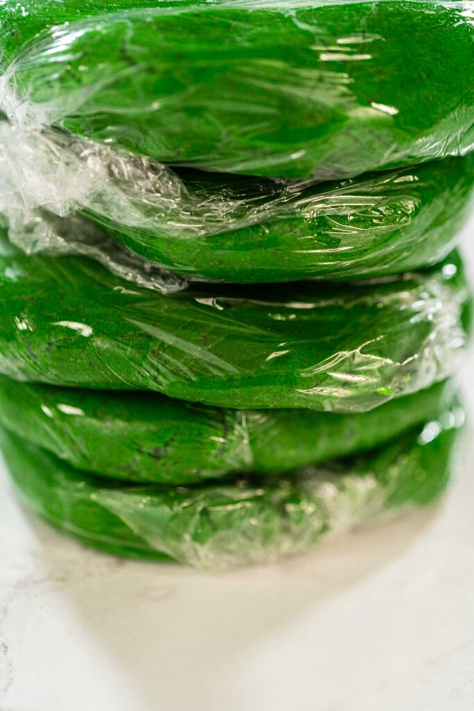 Stacked portions of green dough, each wrapped in plastic wrap, prepared for chilling in the fridge for Vibrant Green Gingerbread Cookies for Festive Baking.