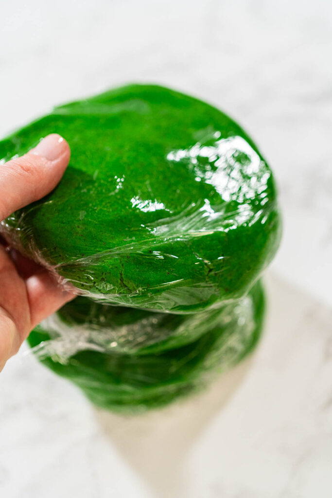 Stacked portions of green dough, each wrapped in plastic wrap, prepared for chilling in the fridge for Vibrant Green Gingerbread Cookies for Festive Baking.
