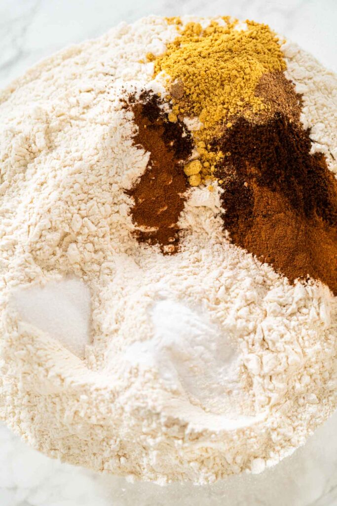 Warm spices like ground cinnamon, cloves, and ginger added to the flour in a glass bowl. These spices are key to achieving the classic gingerbread flavor in the Classic Red Gingerbread Dough.