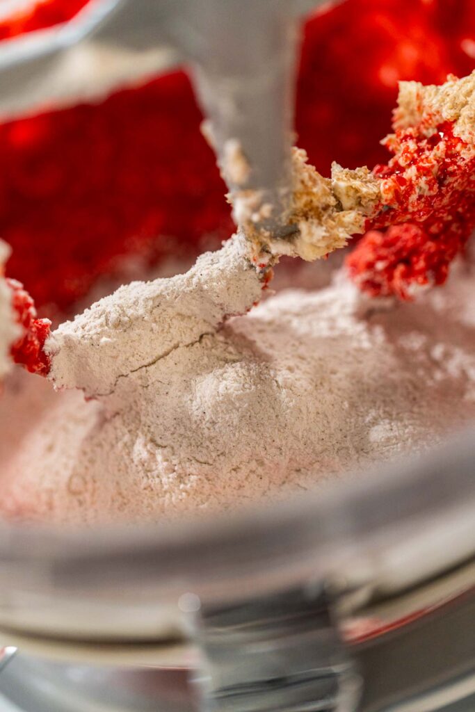Flour mixture is gradually added to the red batter in the mixing bowl. This step thickens the dough while maintaining its bold red color and rich, spiced aroma for the Classic Red Gingerbread Dough.