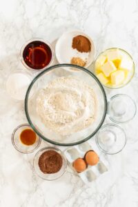 Top view of ingredients for Chocolate Ginger Sugar Cookie Dough on a marble surface, including flour, butter, eggs, cocoa powder, brown sugar, maple syrup, and various spices.