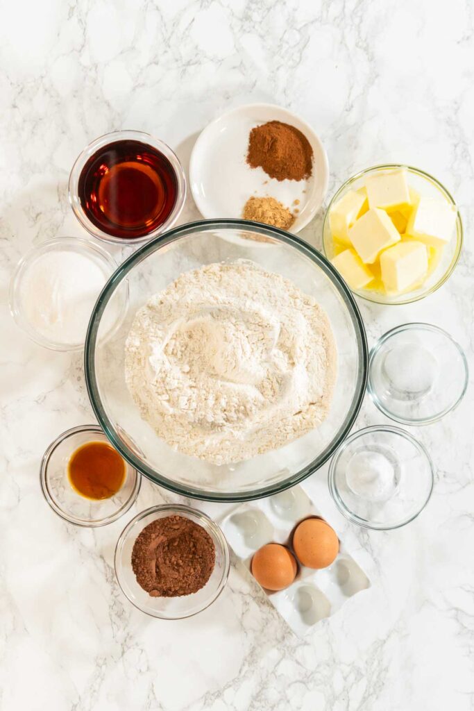 Top view of ingredients for Chocolate Ginger Sugar Cookie Dough on a marble surface, including flour, butter, eggs, cocoa powder, brown sugar, maple syrup, and various spices.