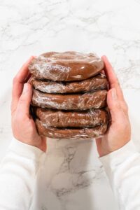 Top view of a stack of Chocolate Ginger Sugar Cookie Dough discs, each wrapped in plastic, held by both hands, showing readiness for storage or chilling.