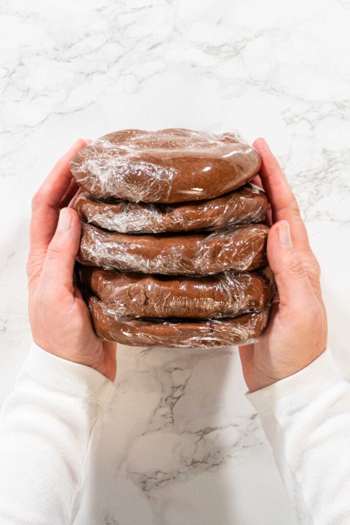 Top view of a stack of Chocolate Ginger Sugar Cookie Dough discs, each wrapped in plastic, held by both hands, showing readiness for storage or chilling.