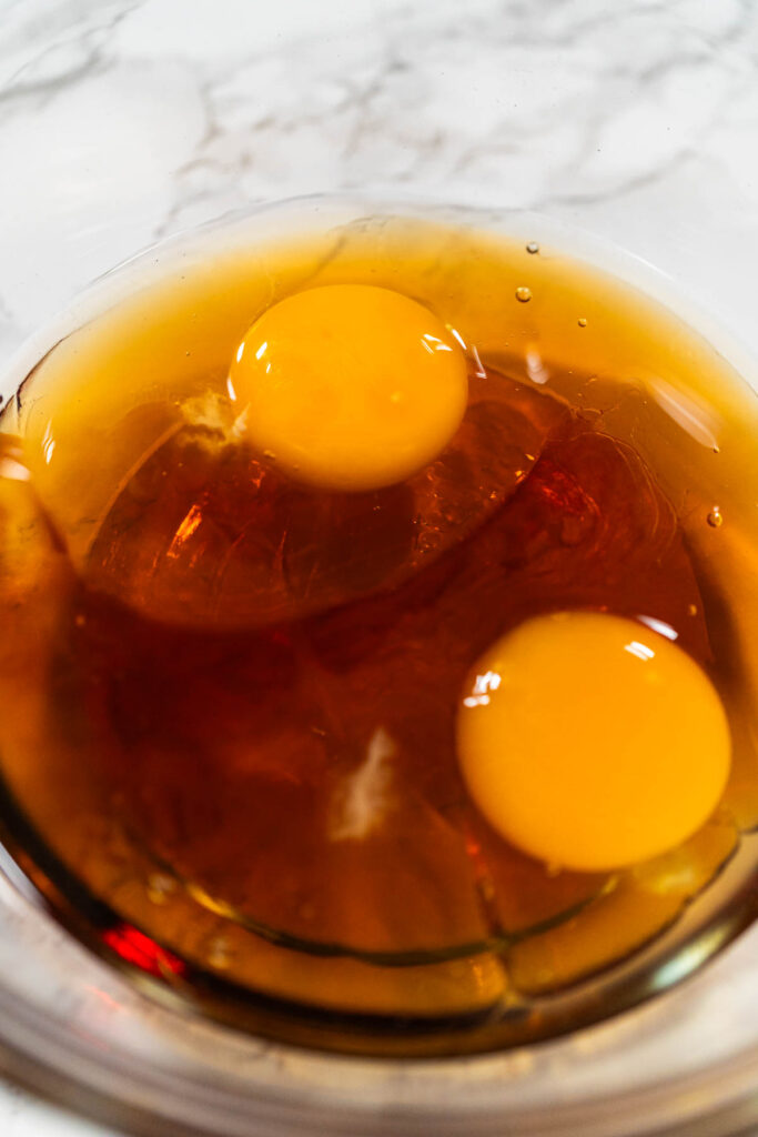 Close-up view of a glass bowl containing two eggs and maple syrup for the Chocolate Ginger Sugar Cookie Dough, creating the wet mixture base.