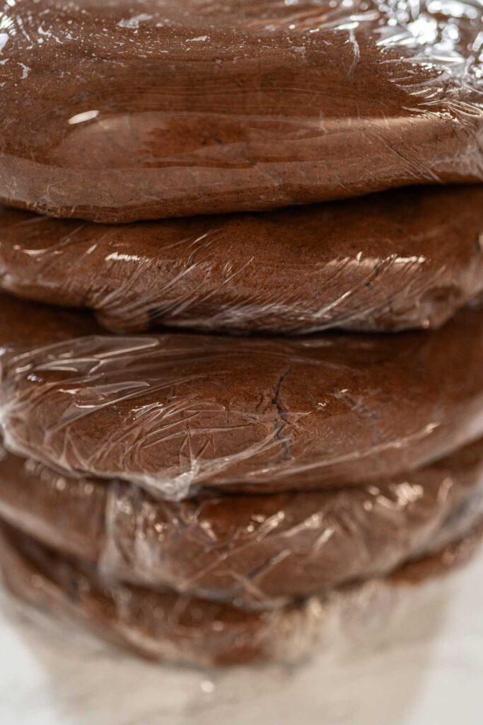 Stacked discs of Chocolate Ginger Sugar Cookie Dough wrapped in plastic, ready to chill, showing a smooth and uniform dough texture.