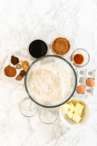 Top-down view of ingredients for Spiced Gingerbread Cookie Dough Perfect for Baking arranged on a marble surface. The central mixing bowl is surrounded by butter, eggs, spices, brown sugar, molasses, and vanilla.