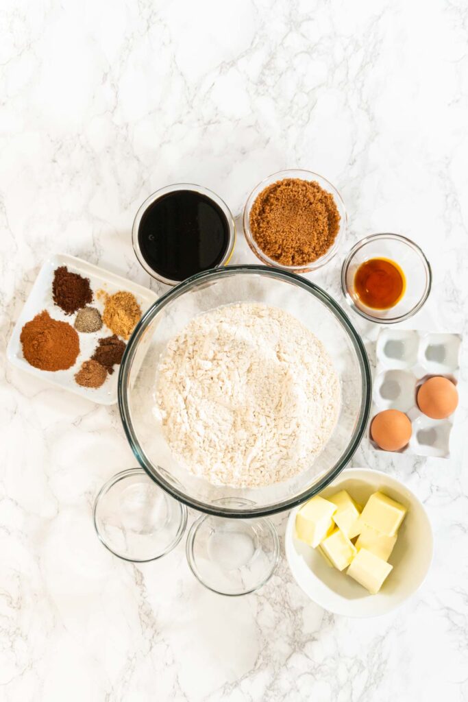 Top-down view of ingredients for Spiced Gingerbread Cookie Dough Perfect for Baking arranged on a marble surface. The central mixing bowl is surrounded by butter, eggs, spices, brown sugar, molasses, and vanilla.