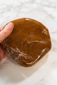 Close-up of a single portion of Spiced Gingerbread Cookie Dough Perfect for Baking wrapped in plastic wrap, held in hand. The dough is prepped and ready for chilling to enhance flavor and texture.