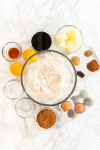 A top-down view showcasing all ingredients for the Zesty Orange Gingerbread Cookie Dough recipe, arranged around a central bowl of flour on a marble countertop. Key ingredients like butter, eggs, cocoa powder, spices, dark syrup, oranges, and small bowls of additional ingredients are visible, ready for preparation.