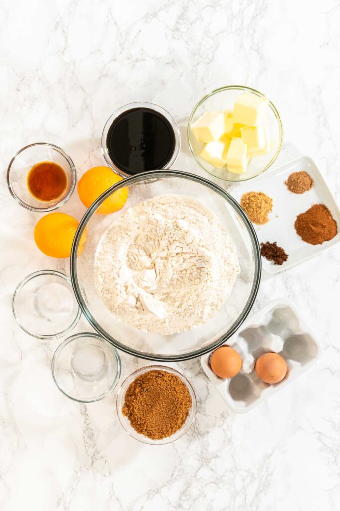 A top-down view showcasing all ingredients for the Zesty Orange Gingerbread Cookie Dough recipe, arranged around a central bowl of flour on a marble countertop. Key ingredients like butter, eggs, cocoa powder, spices, dark syrup, oranges, and small bowls of additional ingredients are visible, ready for preparation.