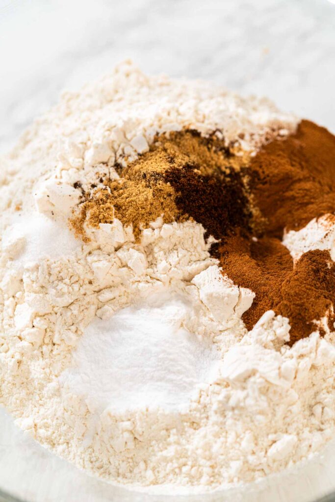 A close-up of dry ingredients for Zesty Orange Gingerbread Cookie Dough, featuring flour, cinnamon, ginger, and cloves in a glass bowl. These spices give the gingerbread its warm and aromatic profile.