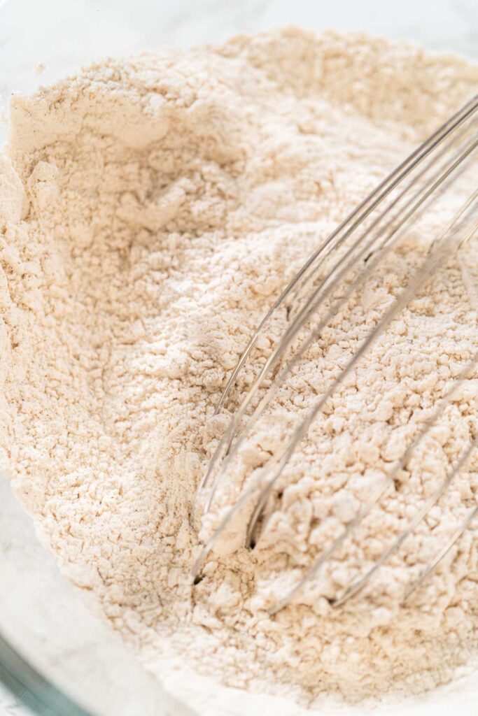 A whisk blending the dry ingredients for Zesty Orange Gingerbread Cookie Dough in a glass bowl, ensuring the spices and flour are evenly combined before adding the wet ingredients.