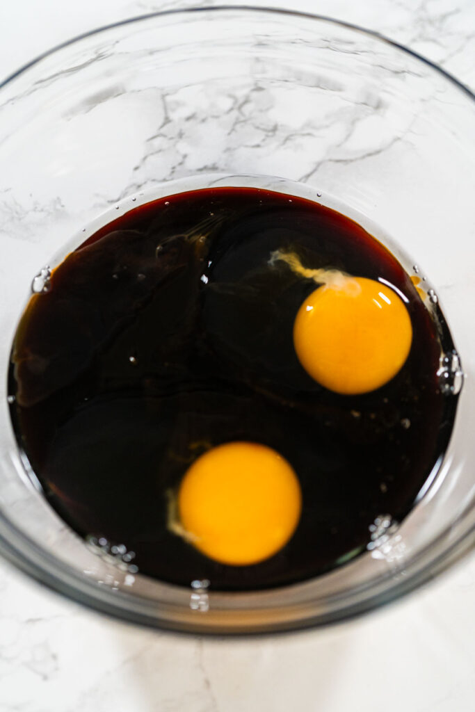 A top-down view of dark molasses and two eggs in a glass bowl, part of the wet ingredients for Zesty Orange Gingerbread Cookie Dough. The molasses adds depth and a rich color to the gingerbread.