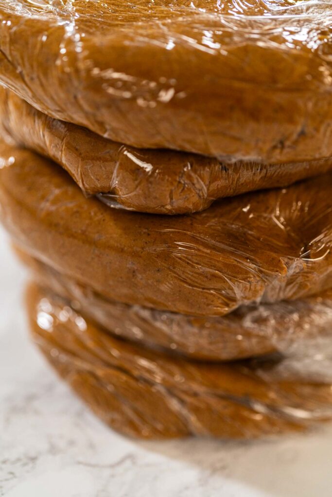 Portions of Zesty Orange Gingerbread Cookie Dough wrapped in plastic wrap and stacked, ready for chilling. Dividing the dough ensures easier handling when rolling out and cutting shapes later.