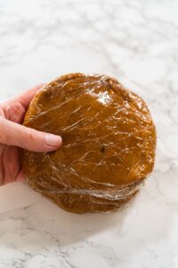 Close-up of a single portion of Zesty Orange Gingerbread Cookie Dough wrapped in plastic wrap, ready to be chilled. The smooth dough surface hints at a rich, flavorful result after baking.