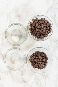 Ingredients Overhead View Description: An overhead view of the main ingredients for Chocolate Royal Icing, featuring semi-sweet chocolate chips and an empty mixing bowl, laid out on a white marble surface.