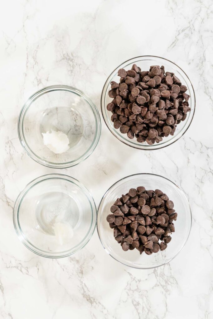 Ingredients Overhead View Description: An overhead view of the main ingredients for Chocolate Royal Icing, featuring semi-sweet chocolate chips and an empty mixing bowl, laid out on a white marble surface.
