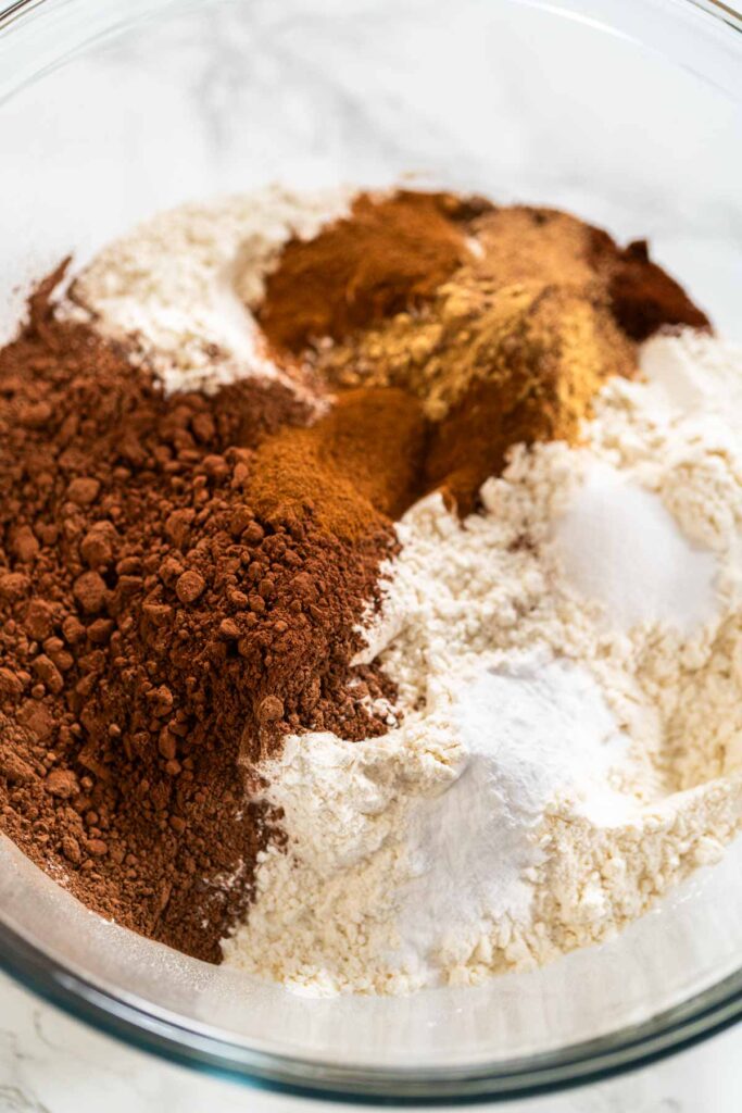 Mixing dry ingredients for Chocolate Gingerbread Cookie Dough, including flour, cocoa powder, cinnamon, ground ginger, and cloves in a glass bowl. The ingredients are layered and ready to be combined, creating a rich, aromatic base for the cookie dough.