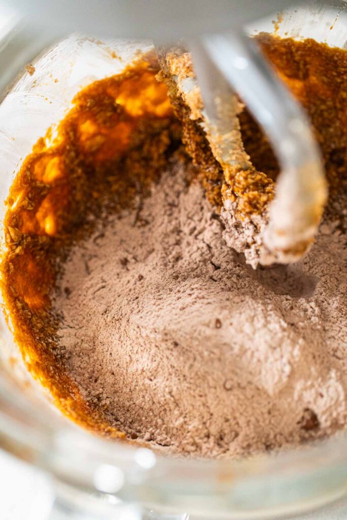 Adding dry ingredients, including flour and cocoa powder, to the wet ingredients in the mixer for Chocolate Gingerbread Cookie Dough. This step begins the process of forming a soft and flavorful dough.