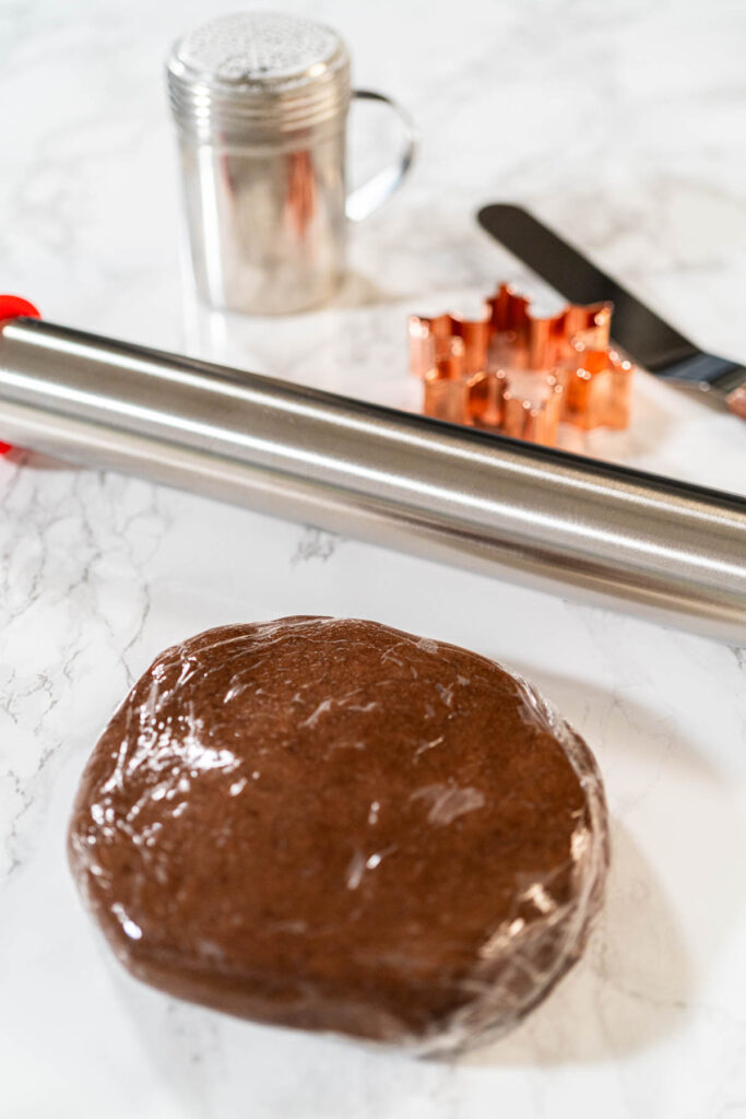 Chocolate Ginger Sugar Cookies in Snowflake Shapes A Festive Tre - Arina Photography A ball of Chocolate Ginger Sugar Cookie dough wrapped in plastic, placed on a marble surface with baking tools in the background. Close-up perspective.