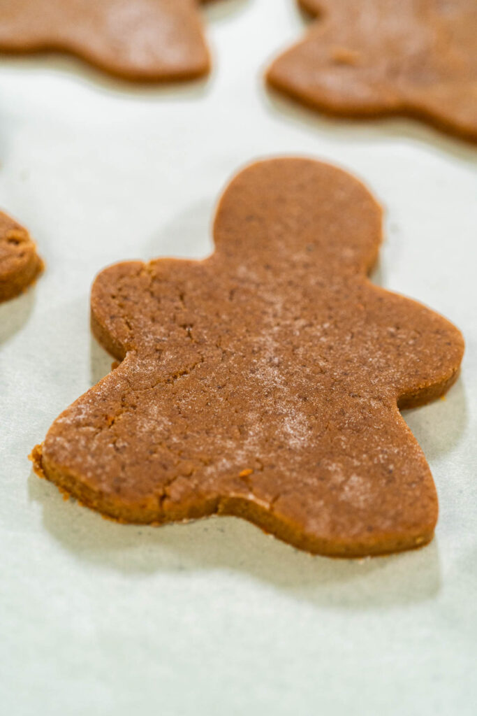 A tray of freshly baked Orange Zest Gingerbread Cookies in gingerbread girl shapes cooling on parchment paper. The close-up perspective highlights their golden-brown texture.