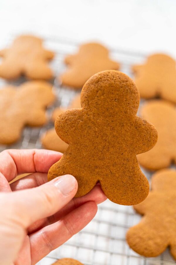 Close-up of a hand holding a baked gingerbread man cookie over a cooling rack filled with cookies.