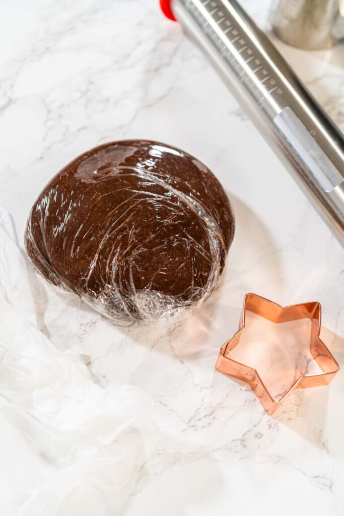 Chocolate-spiced gingerbread cookie dough wrapped in plastic wrap with a copper star-shaped cookie cutter on a marble surface.
