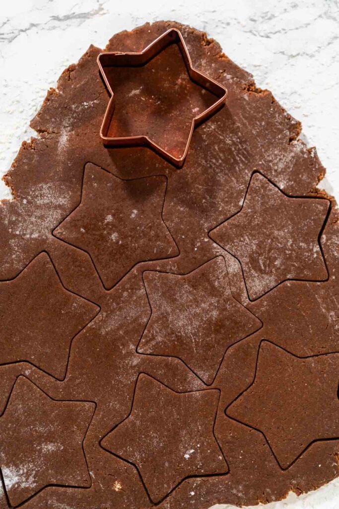 Star-shaped cookie cutter cutting out shapes from rolled chocolate-spiced gingerbread dough on a floured surface.